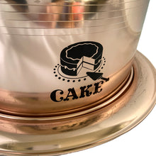 Load image into Gallery viewer, Vintage copper/rose gold toned aluminum dome cake carrier keeper featuring a black bakelite handle and an illustration in black of a sliced cake with the word "CAKE" below in a cute vintage font and locking levers to hold the dome securely to the base. The perfect vessel to store your home-baked or store bought confections! In as found vintage condition with wear to the black at the top of the dome, age-related wear. Fits up to a 10 1/2 x 5 inch cake Overall measures 14 1/2 x 8 inches