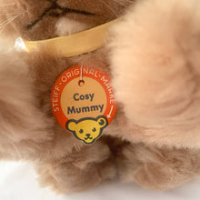 Load image into Gallery viewer, Vintage stuffed animal tan light brown bunny rabbit. Washable Dralon plush material. Hand embroidered pink nose and brown mouth, brown glass eyes with black pupils, and light beige felt rear foot pads. Gold satin ribbon and round red, blue and yellow tag bearing the name Cosy Mummy. Brass button-in-ear yellow and black tag 494020 and paper tag. Steiff, Germany 1970s. This bunny rabbit sits in a seated or reclining position. In excellent condition. Measures 8 1/2 inches or 22cm