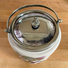 Load image into Gallery viewer, Antique porcelain biscuit jar featuring whimsical florals on a cream ground with silver plated collar, handle and lid. Crafted by Taylor Tunnicliffe, England, 1875 - 1898. Absolutely stunning example of decorative and functional artistry from the Aesthetic Movement era! In overall excellent condition for a piece of its age with wear to the silver plate on the lid and minor discolouration on the porcelain. Measures 5 x 6 5/8 inches (8 1/4 with handle raised)