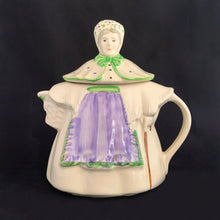 Load image into Gallery viewer, Vintage cream coloured figural "Granny Ann" ceramic teapot featuring a sweet granny holding a brown cane and dressed in a hat and garden frock in green and purple. Crafted by Shawnee Pottery, USA, circa 1940s. Even the spout is a figural basket! In excellent condition, free from chips/cracks/repairs. Marked on the bottom, "Patented Granny Ann U.S.A." Measures 8 x 5 5/8 x 8 1/2 inches
