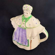 Load image into Gallery viewer, Vintage cream coloured figural "Granny Ann" ceramic teapot featuring a sweet granny holding a brown cane and dressed in a hat and garden frock in green and purple. Crafted by Shawnee Pottery, USA, circa 1940s. Even the spout is a figural basket! In excellent condition, free from chips/cracks/repairs. Marked on the bottom, "Patented Granny Ann U.S.A." Measures 8 x 5 5/8 x 8 1/2 inches
