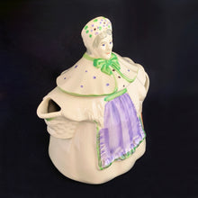 Load image into Gallery viewer, Vintage cream coloured figural "Granny Ann" ceramic teapot featuring a sweet granny holding a brown cane and dressed in a hat and garden frock in green and purple. Crafted by Shawnee Pottery, USA, circa 1940s. Even the spout is a figural basket! In excellent condition, free from chips/cracks/repairs. Marked on the bottom, "Patented Granny Ann U.S.A." Measures 8 x 5 5/8 x 8 1/2 inches