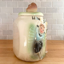 Load image into Gallery viewer, This rare vintage ceramic corner cookie jar features brunette and blonde cheerleaders in old-fashioned outfits, cheering underneath the words Have A Cookie in between is a megaphone on the grass that reads cookie time all glazed in shades of green, blue, yellow brown and black on a cream ground, topped with a rust-brown megaphone on the lid. The piece-de-resistance are the cheerleaders holographic facial expressions that change from eyes open with a wide smile to eyes shut with closed smiles.
