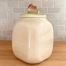 Load image into Gallery viewer, This rare vintage ceramic corner cookie jar features brunette and blonde cheerleaders in old-fashioned outfits, cheering underneath the words Have A Cookie in between is a megaphone on the grass that reads cookie time all glazed in shades of green, blue, yellow brown and black on a cream ground, topped with a rust-brown megaphone on the lid. The piece-de-resistance are the cheerleaders holographic facial expressions that change from eyes open with a wide smile to eyes shut with closed smiles.