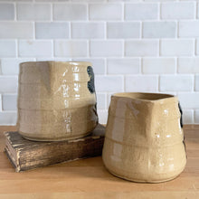 Load image into Gallery viewer, Vintage natural yellow ware strapped barrel milk pitcher featuring navy lily in bas relief with rope style handle, No. 21171. Crafted by Robinson Ransbottom Pottery Co., USA, 1930s. Enhance your home's decor with this beautiful piece of primitive farmhouse tableware. A wonderful addition to any art pottery collection!
Excellent vintage condition, no chips/cracks/repairs, age-related crazing and typical manufacturer's glaze pops. Unmarked
Option 1: 4 x 4 1/8 inches. 32 oz
Option 2: 5 1/4 x 5 1/4. 48 oz