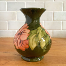 Load image into Gallery viewer, Vintage art pottery vase featuring tubelined hibiscus flowers hand painted in shades of peach on an avocado green ground. Design by Walter Moorcroft. Crafted by Moorcroft Pottery, England, 1980s. A beautiful piece for your art pottery collection or to give as a gift! In excellent condition, no chips/cracks/repairs. Bottom has impressed marks, "MOORCROFT POTTERY" AND "MADE IN ENGLAND" and gold oval MOORCROFT, England sticker. Measures 3 3/4 x 5 1/4 inches