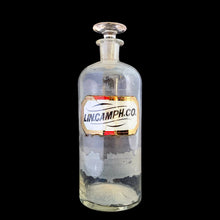 Load image into Gallery viewer, Late 19th century antique hand blown glass pharmacy chemist apothecary jar with stopper. The hand painted label reads LIN.CAMPH.CO. and the label glass has a gold border. Pontil scar on the bottom. These bottles were crafted by skilled glassblowers and used to house chemical compounds in pharmacies...a cool piece of history. In as found condition, there appears to be a crack at the neck and the glass over the label has damage, see photos. Measures 3 3/8 x 9 5/8 inches