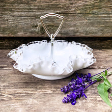 Load image into Gallery viewer, This vintage milk glass "Silver Crest" bon bon dish brings all the drama in a small package! It features a clear crimped edge over milk glass and a silver-toned metal handle. Crafted by the Fenton Art Glass, USA, circa 1960s. A graceful piece of pressed glass in a classic pattern that will always be in style no matter your decor style! Fill with your favourite candies, nuts or snacks. In excellent condition, free form chips/cracks. Measures 8 x 5 inches (incl. handle)