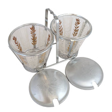 Load image into Gallery viewer, Vintage 2-part glass condiment server featuring alternating 22kt gold leaves and clear leaves on a white stripe and pinecone embossed aluminum lids in chrome caddy. Crafted by Dominion Glass, Canada, 1950s. Love the conical shape of the glass and the styling on the caddy....very mid-century modern and a great way to serve your favourite fixings.
In excellent condition, no chips.
Overall measures 7 3/4 x 4 x 5 1/2 inches