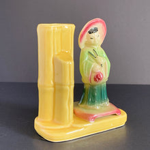 Load image into Gallery viewer, A vintage ceramic vase of planter in the Chinoiserie style features a green and red Asian figure with a yellow bamboo vase on a podium, shape 702. Crafted by Shawnee Pottery USA. Perfect as a bud vase or to a decor accent.
In good vintage condition, no cracks. Small glaze miss on the front of the podium.
Measures 5 x 2 1/2 x 5 1/2 inches