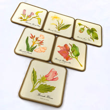 Load image into Gallery viewer, A vintage set of six square round edged coasters, each decorated with an illustration of a tropical flower on an off-white background, bordered in gold with cork-backing. Coasters make the perfect resting place for drinking glasses or mugs to protect your furniture from being marked. Made by Pimpernel in England. Makes a great housewarming or hostess gift! In excellent vintage condition. Each coaster measures 4-1/8" x 4-1/8"