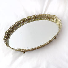 Load image into Gallery viewer, This elegant vintage Hollywood Regency style oval-shaped mirrored dresser vanity tray, featuring brass ormolu details, is perfect for displaying perfume bottles, trinket dishes, and makeup. Alternately, hang it on the wall to add glitz and glamour to your décor. In excellent condition. Measures 12 x 9 x 1 1/4 inches