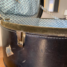 Load image into Gallery viewer, black vintage round train case or suitcase with nice blue and white floral/diamond patterned lining, a leather handle, brass latches and locking mechanism with TWO original keys. Exterior has two stickers, HAPAG shipping company and a customs inspection sticker. Well worn leather handle. All hardware is in good working order and the case opens and closes nicely. The case has normal wear from 80 plus years of usage, but no major damage. measures 16-1/2" tall and 18" wide, and 9" deep.