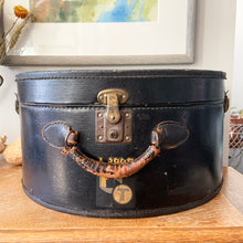 Load image into Gallery viewer, black vintage round train case or suitcase with nice blue and white floral/diamond patterned lining, a leather handle, brass latches and locking mechanism with TWO original keys. Exterior has two stickers, HAPAG shipping company and a customs inspection sticker. Well worn leather handle. All hardware is in good working order and the case opens and closes nicely. The case has normal wear from 80 plus years of usage, but no major damage. measures 16-1/2" tall and 18" wide, and 9" deep.