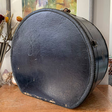Load image into Gallery viewer, black vintage round train case or suitcase with nice blue and white floral/diamond patterned lining, a leather handle, brass latches and locking mechanism with TWO original keys. Exterior has two stickers, HAPAG shipping company and a customs inspection sticker. Well worn leather handle. All hardware is in good working order and the case opens and closes nicely. The case has normal wear from 80 plus years of usage, but no major damage. measures 16-1/2" tall and 18" wide, and 9" deep.