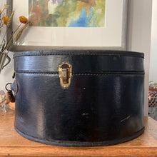 Load image into Gallery viewer, black vintage round train case or suitcase with nice blue and white floral/diamond patterned lining, a leather handle, brass latches and locking mechanism with TWO original keys. Exterior has two stickers, HAPAG shipping company and a customs inspection sticker. Well worn leather handle. All hardware is in good working order and the case opens and closes nicely. The case has normal wear from 80 plus years of usage, but no major damage. measures 16-1/2" tall and 18" wide, and 9" deep.