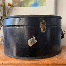Load image into Gallery viewer, black vintage round train case or suitcase with nice blue and white floral/diamond patterned lining, a leather handle, brass latches and locking mechanism with TWO original keys. Exterior has two stickers, HAPAG shipping company and a customs inspection sticker. Well worn leather handle. All hardware is in good working order and the case opens and closes nicely. The case has normal wear from 80 plus years of usage, but no major damage. measures 16-1/2" tall and 18" wide, and 9" deep.