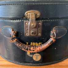 Load image into Gallery viewer, black vintage round train case or suitcase with nice blue and white floral/diamond patterned lining, a leather handle, brass latches and locking mechanism with TWO original keys. Exterior has two stickers, HAPAG shipping company and a customs inspection sticker. Well worn leather handle. All hardware is in good working order and the case opens and closes nicely. The case has normal wear from 80 plus years of usage, but no major damage. measures 16-1/2" tall and 18" wide, and 9" deep.