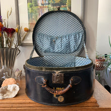 Load image into Gallery viewer, black vintage round train case or suitcase with nice blue and white floral/diamond patterned lining, a leather handle, brass latches and locking mechanism with TWO original keys. Exterior has two stickers, HAPAG shipping company and a customs inspection sticker. Well worn leather handle. All hardware is in good working order and the case opens and closes nicely. The case has normal wear from 80 plus years of usage, but no major damage. measures 16-1/2" tall and 18" wide, and 9" deep.