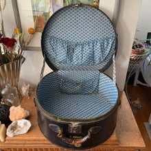 Load image into Gallery viewer, black vintage round train case or suitcase with nice blue and white floral/diamond patterned lining, a leather handle, brass latches and locking mechanism with TWO original keys. Exterior has two stickers, HAPAG shipping company and a customs inspection sticker. Well worn leather handle. All hardware is in good working order and the case opens and closes nicely. The case has normal wear from 80 plus years of usage, but no major damage. measures 16-1/2" tall and 18" wide, and 9" deep.