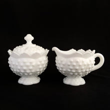 Load image into Gallery viewer, Vintage white milk glass is the epitome of simple elegance! This is a scalloped creamer and lidded sugar bowl in the "Hobnail" pattern. Produced by Fenton, post 1970. Marked. The creamer is in excellent condition, free from chips or cracks. The sugar bowl has a chip on the side of one of the scalloped points...it's not rough or really noticeable thanks to the piece being white. Creamer measures 4" 3-1/2", sugar w/ lid measures 4" x 4-3/4"