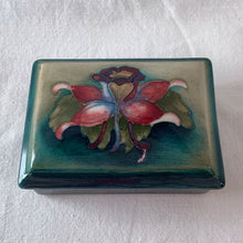 Load image into Gallery viewer, A lovely vintage "Columbine" dresser box with a jewel-toned flower against a green wash ground. Design by Walter Moorcroft and produced by Moorcroft Pottery, England, circa 1950. Stamped with the WM Moorcroft signature and "Made in England" along with paper label having the Royal Warrant and "By Appointment, Potter to the late Queen Mary". In excellent condition, free from chips, cracks, repairs; crazing present. Measures 4.75" x 3.5" x 2"