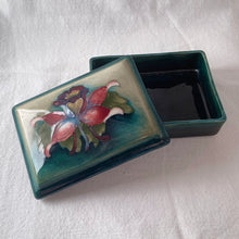 Load image into Gallery viewer, A lovely vintage "Columbine" dresser box with a jewel-toned flower against a green wash ground. Design by Walter Moorcroft and produced by Moorcroft Pottery, England, circa 1950. Stamped with the WM Moorcroft signature and "Made in England" along with paper label having the Royal Warrant and "By Appointment, Potter to the late Queen Mary". In excellent condition, free from chips, cracks, repairs; crazing present. Measures 4.75" x 3.5" x 2"