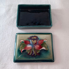 Load image into Gallery viewer, A lovely vintage "Columbine" dresser box with a jewel-toned flower against a green wash ground. Design by Walter Moorcroft and produced by Moorcroft Pottery, England, circa 1950. Stamped with the WM Moorcroft signature and "Made in England" along with paper label having the Royal Warrant and "By Appointment, Potter to the late Queen Mary". In excellent condition, free from chips, cracks, repairs; crazing present. Measures 4.75" x 3.5" x 2"