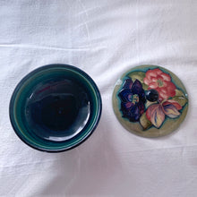 Load image into Gallery viewer, Vintage mid-century "Clematis" lidded dish with green ground lid and teal/blue ground on bowl's interior and deep blue on the exterior of the bowl. The colours are vibrant and the tube lining and hand painting are spectacular. Produced by Moorcroft Pottery, England, circa 1950s.  Measures 6-1/4" x 3-1/2"  Excellent condition. No cracks or chips. Normal crazing for a piece of this age. Impressed marks include Walter Moorcroft's signature, Potter to H.M. The Queen and Made in England.