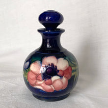 Load image into Gallery viewer, Vintage mid-century "Anemone" perfume bottle with stopper. Hand painted in the slip glaze technique in shades of purple, pink and green on a deep cobalt blue ground. Produced by Moorcroft Pottery, circa 1928-1953. In excellent condition, no chips, cracks or repairs, crazing present. Bottom shows impressed signature of Walter Moorcroft, "MADE IN ENGLAND" and Royal Warrant paper label - 'Potter To HM The Queen'. Measures 4" x 5-1/2"