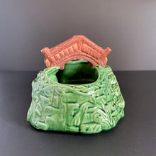 Load image into Gallery viewer, A vintage ceramic planter featuring a rusty red bridge over a green pond with impressed brick wall and stairs, and floral details. Crafted by Shawnee Pottery, USA, 1950s. Create a unique garden with this unique planter!
In vintage condition with minor flea bites to the bridge.
Measures 9 x 5 3/4 x 4 3/4 inches