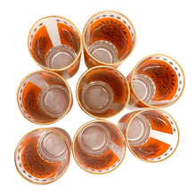 Load image into Gallery viewer, Overhead view of eight vintage 1960s Jeannette Glass Celestial Aztec Sun highball glasses showing orange geometric sunburst patterns with 22K gold accents
