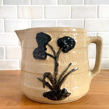 Load image into Gallery viewer, Vintage natural yellow ware strapped barrel milk pitcher featuring black lily in bas relief with rope style handle, No. 21171. Crafted by Robinson Ransbottom Pottery Co., USA, 1930s. A beautiful piece of primitive farmhouse tableware to enhance your home&#39;s decor and a wonderful addition to any art pottery collection!

Measures 5 1/4 x 5 1/4 inches. In as found vintage condition, chip to rim, no cracks/repairs, glaze wear at the spout, age-related crazing and glaze pops. Unmarked. Capacity 48 ounces
