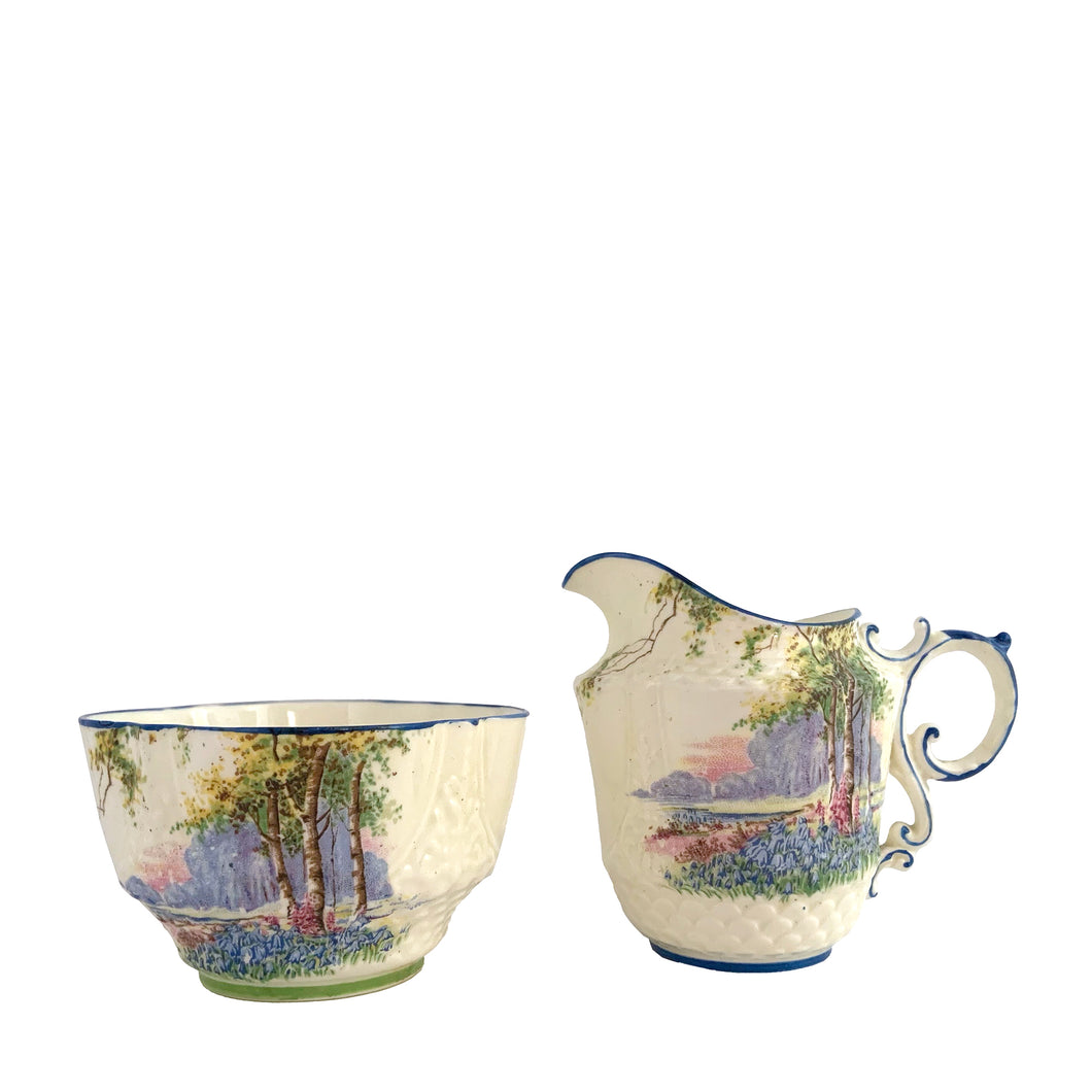 Vintage Bluebell Time bone china creamer and sugar featuring a delicately shaped vessels decorated with springtime woodland scene with bluebell flowers amongst groves of trees and trimmed in blue. Crafted by Aynsley, England, 1934 - 1950s.

In excellent condition, no chips/cracks/repairs. Maker's mark present with Bluebell Time and As supplied to H.M. the Queen.

Creamer measures 2 1/2 x 3 1/8 inches

Sugar measures 3 1/4 x 1 7/8 inches