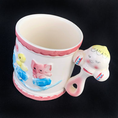 Kitschy cuteness abounds in this vintage mid-century ceramic child's mug featuring a figural baby faced rattle along with an embossed train on its track with a sweet yellow chick and pink elephant in alphabet cars, rimmed in pink scalloped borders at the top and bottom. Crafted by Rubens, Japan, circa 1950s. Such a sweet piece for a nursery and would make a lovely baby shower gift!