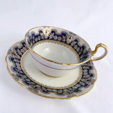 Load image into Gallery viewer, Antique bone china teacup and saucer is absolutely stunning! Both have a subtle scalloped shape. The white cup is decorated in a design of pale blue floral bouquets and bows on a cobalt blue band with gold gilt details. Produced by Aynsley, England. Excellent condition, free from chips, cracks and repairs. The pattern number 3410 is painted on the saucer. The stamped maker&#39;s mark indicates production between 1905 - 1925. Teacup measures 3 1/2 x 2 1/4 inches | Saucer measures 5 5/8 inches
