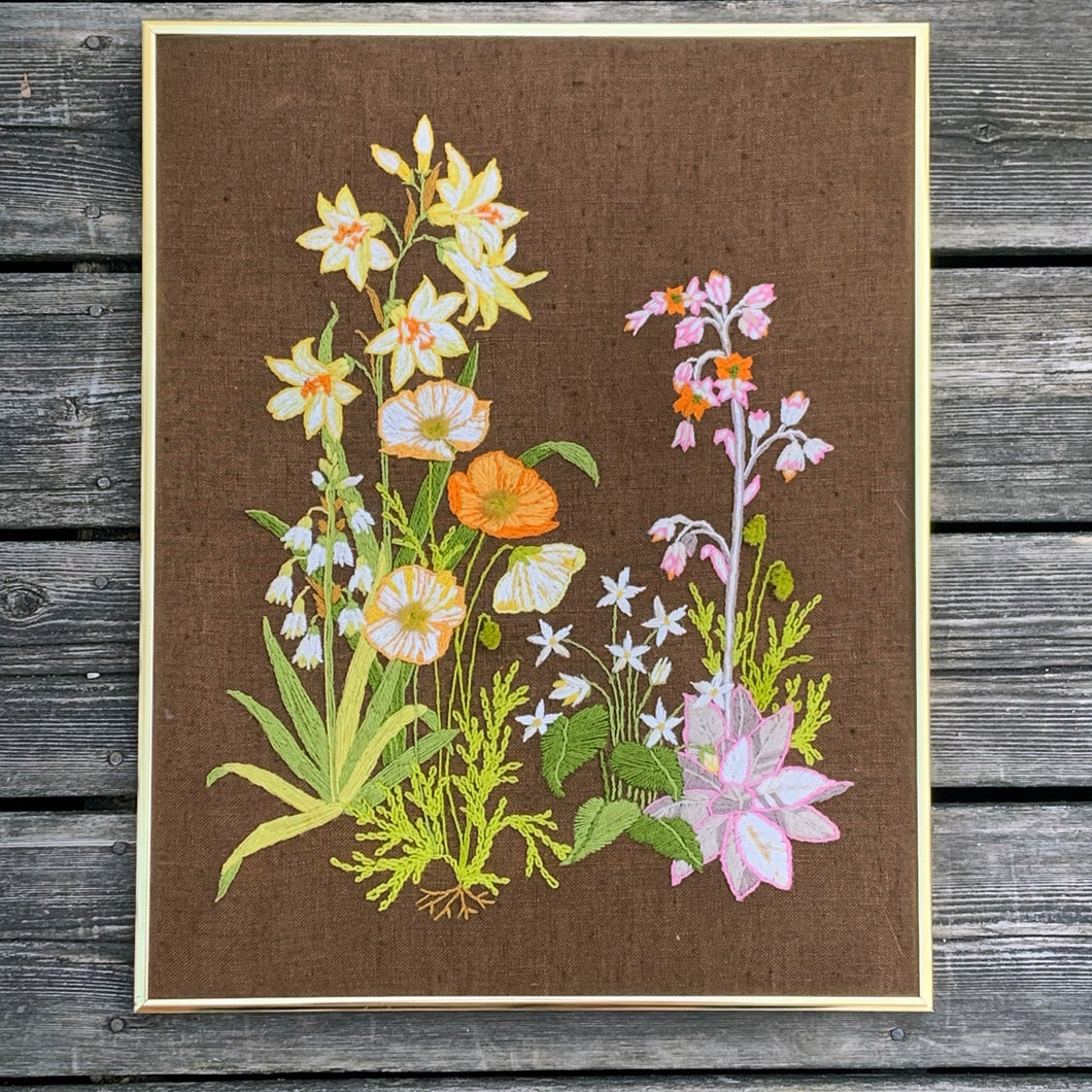 Vintage hand stitched crewel of a floral still life in shades of orange, yellow, pink and green on deep brown linen which makes the design pop! This piece is frame in gold-toned metal. There is a tag on the back that states, 