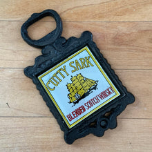 Load image into Gallery viewer, Vintage Cutty Sark Blended Scotch Whiskey Promotional Cast Iron Bottle Opener with back painted glass insert. A fun and practical conversation piece for your cottage or farmhouse barware and decor!

In excellent condition.

Measures 2 3/4 x 4 3/4 inches
