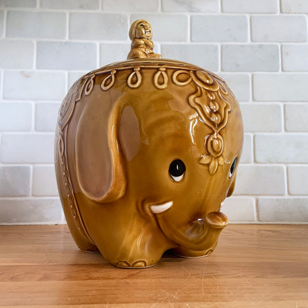 vintage cookie jar of figural elephant features a turbin wearing gentleman sitting on a pillow riding on top which forms the lid's finial. adorable and the expression on the rider's face is sassy! The elephant is beautifully decorated in an East Indian motif. Crafted by Noritake, Japan for Giftcraft export. In outstanding condition, free from chips/cracks/wear. We believe this is new old stock. Original sticker present and the bottom has the stamped maker's mark. Measures 7 1/2 x 5 1/2 x 9 3/4 inches