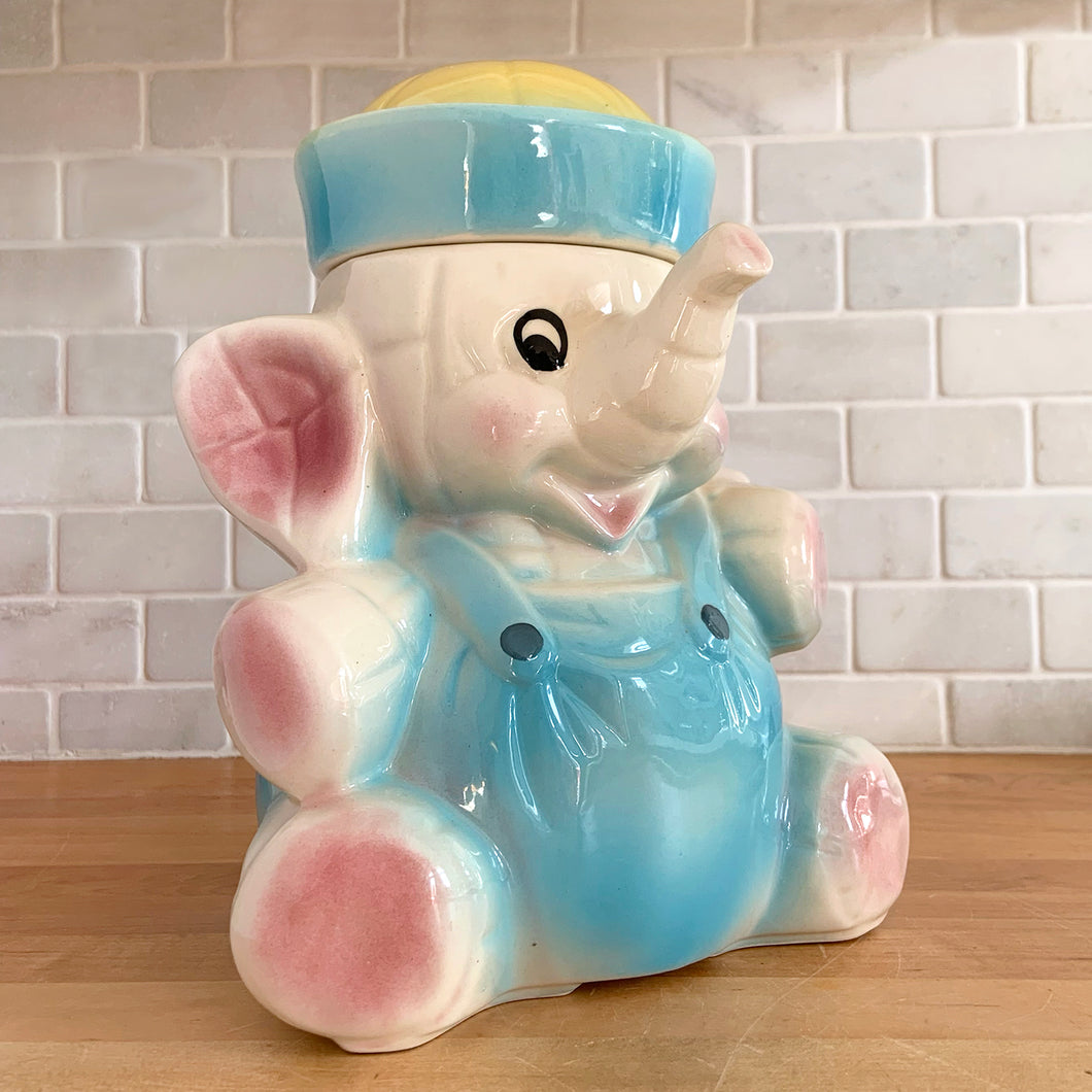 Vintage mid-century hand painted figural white elephant ceramic cookie jar with blue and yellow sailor hat and overalls and pink details. Crafted by American Bisque, USA, circa 1950s. Add some kitsch to your kitchen decor with this adorable collectible!

In excellent condition, no chips or cracks, age-related crazing. Marked USA on the back of the jar.

Measures 8 1/2 x 6 x 10 1/2 inches