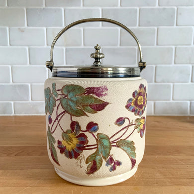 Antique porcelain biscuit jar featuring whimsical florals on a cream ground with silver plated collar, handle and lid. Crafted by Taylor Tunnicliffe, England, 1875 - 1898. Absolutely stunning example of decorative and functional artistry from the Aesthetic Movement era!  In overall excellent condition for a piece of its age with wear to the silver plate on the lid and minor discolouration on the porcelain.  Measures 5 x 6 5/8 inches (8 1/4 with handle raised)
