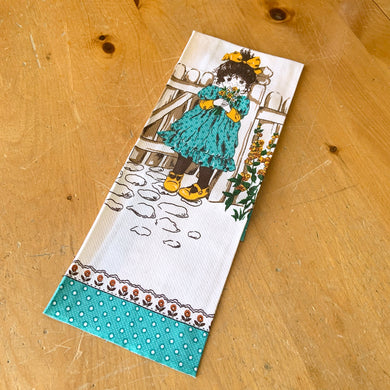 Pretty vintage white cotton tea towel featuring a girl dressed in turquoise and yellow holding flowers beside a garden gate. The towel is bordered in turquoise and red flowers. In excellent condition. Measures 17 x 28 inches