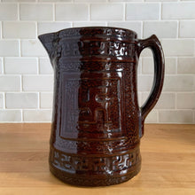 Load image into Gallery viewer, Antique brown glazed stoneware tankard or pitcher, decorated with good luck swastikas. In Native American cultures, the swastika, or Whirling Log as it&#39;s known in Navajo, was a symbol of good fortune, the vehicle of the Culture Hero, or the four winds. This design pre-dates the Nazi era. Crafted by McCoy Pottery, 1920s.

In as found vintage condition, no chips/cracks/repairs. The interior has glaze loss on the bottom and hard water marks.

Measures 7 3/4 x 5 3/4 x 8 1/4 inches (incl. spout/handle)
