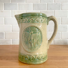 Load image into Gallery viewer, Antique grazing cows salt glazed yellow ware ceramic pitcher with decorative green medallion, rim and base. Crafted by Hull Pottery, USA, 1920s. This utilitarian piece nicely complement any farmhouse or cottage kitchen!

In as found vintage condition, no chips/repairs, hairline crack near the spout (see photos), age-related wear to the exterior. Unmarked.

Measures 6 1/2 x 6 1/2 inches (incl. handle)

Capacity approximately 32 ounces
