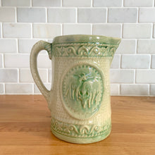 Load image into Gallery viewer, Antique grazing cows salt glazed yellow ware ceramic pitcher with decorative green medallion, rim and base. Crafted by Hull Pottery, USA, 1920s. This utilitarian piece nicely complement any farmhouse or cottage kitchen!

In as found vintage condition, no chips/repairs, hairline crack near the spout (see photos), age-related wear to the exterior. Unmarked.

Measures 6 1/2 x 6 1/2 inches (incl. handle)

Capacity approximately 32 ounces
