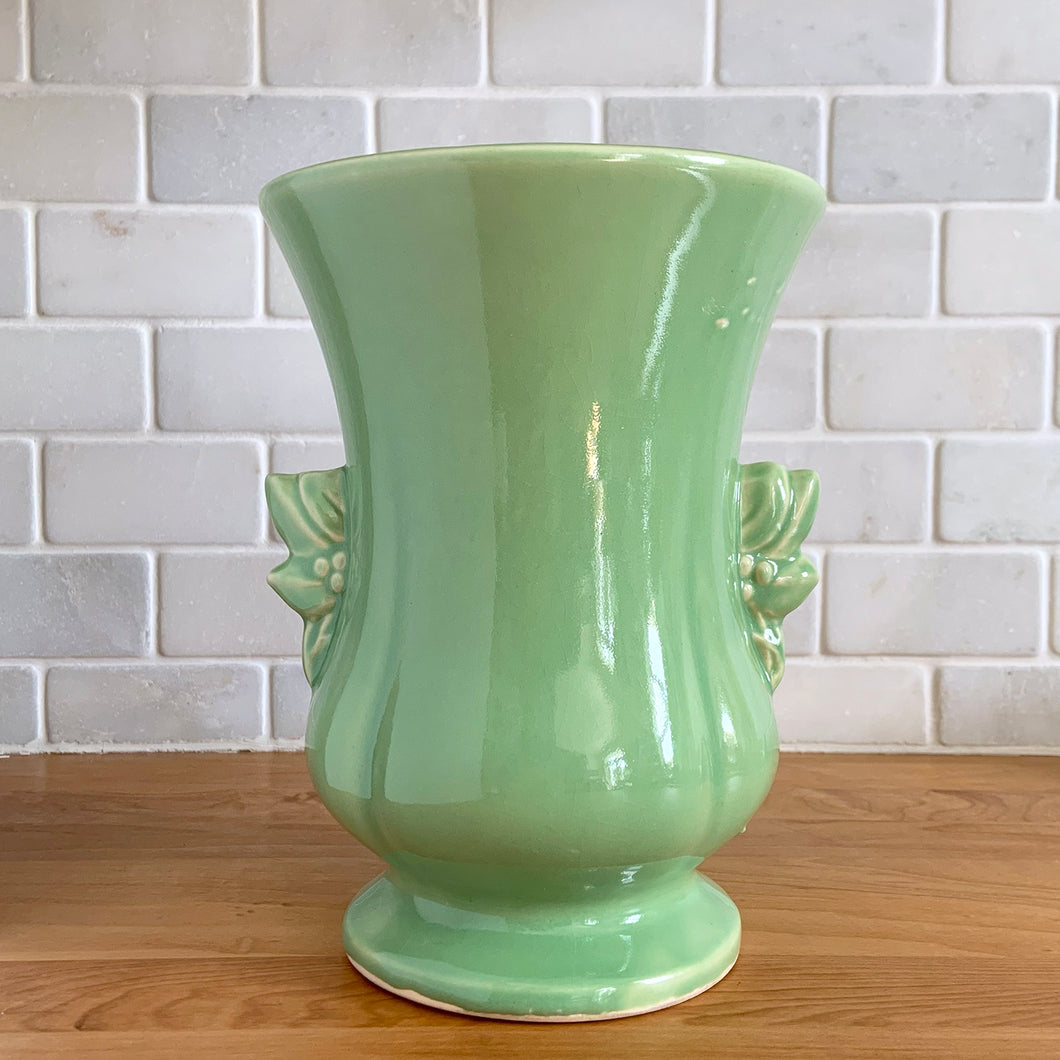 Beautiful vintage Art Deco pale green handled trumpet vase #282 features berry and leaf handles. Crafted by McCoy Pottery, 1946. This vase is must for any art pottery collector. A lovely way to enhance your home's decor or to give as a gift!

In great vintage condition, chip to underside of foot, no cracks/repairs. Typical manufacturer's glaze pops/defects. Maker's mark on the bottom.

Measures 6 x 8 1/8 inches (incl. handle/spout)