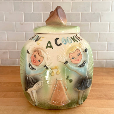 This rare vintage ceramic corner cookie jar features brunette and blonde cheerleaders in old-fashioned outfits, cheering underneath the words Have A Cookie in between is a megaphone on the grass that reads cookie time all glazed in shades of green, blue, yellow brown and black on a cream ground, topped with a rust-brown megaphone on the lid. The piece-de-resistance are the cheerleaders holographic facial expressions that change from eyes open with a wide smile to eyes shut with closed smiles. 