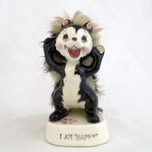 Load image into Gallery viewer, A vintage-style ceramic figurine of a black and white skunk standing on a white base that reads 'I AM HAPPY'. The skunk has pink inner ears, a red nose and mouth, and fluffy beige and brown hair or fur details.
