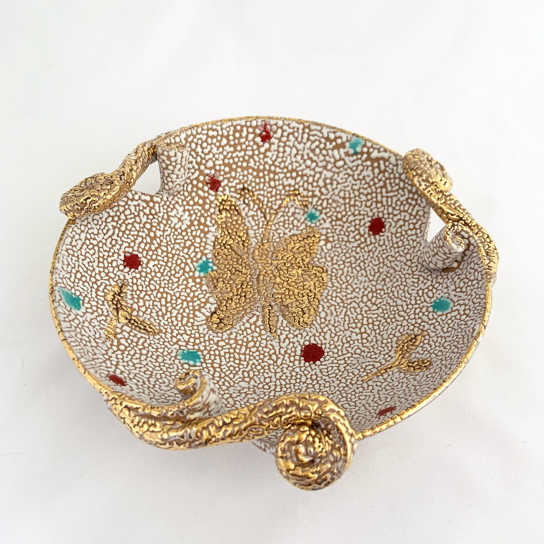 A fabulous piece of Bitossi mid-century lava pebble glazed pottery dish featuring a gold butterfly and three decorative scrolled and curled handles. Artisan crafted in Italy, and imported to North America by Irving Richards of Raymor, USA, circa 1960s. Makes a lovely trinket or candy dish. A phenomenal gift for the MCM lover. Excellent condition, free from chips/cracks/repairs. Marked Italy. Measures 6 1/2 x 2 3/8 inches