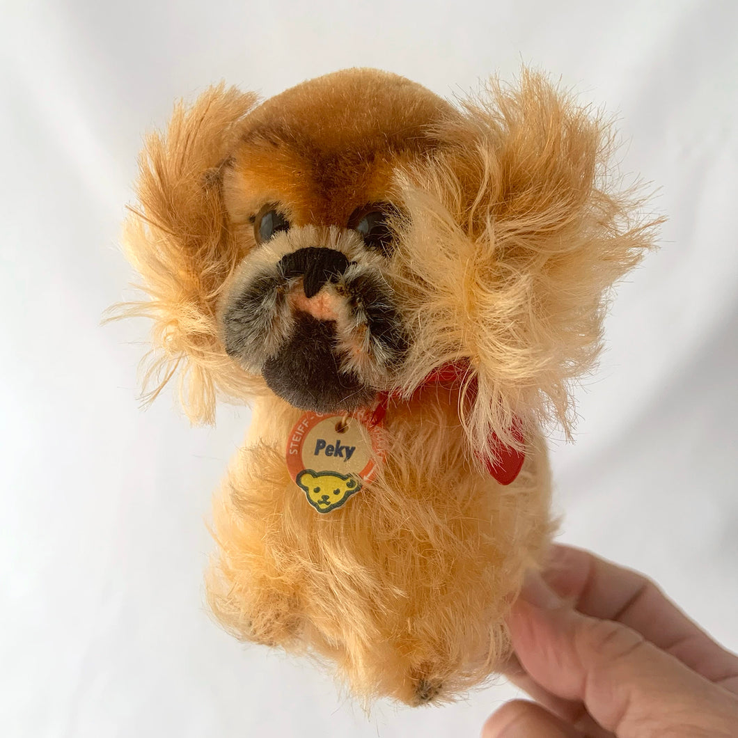 Vintage Peky Pekingese dog stuffed animal features orange amber mohair. He has a jointed head, hand embroidered black nose and mouth, brown glass eyes with black pupils. On its neck is a red ribbon with the original round red, blue and yellow tag bearing the name Peky. Ear has Steiff's silver script button, with remnants of the yellow tag. Crafted by Steiff, Germany 1959 - 1964. A must-have for any Steiff or stuffie collector! In excellent condition. Measures 4 inches or 10cm tall